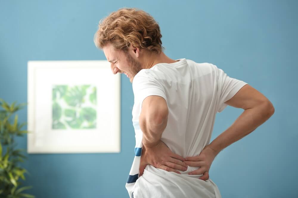 Junger Mann greift sich mit schmerzverzerrtem Gesicht bei Rückenschmerzen in den mittleren Rücken