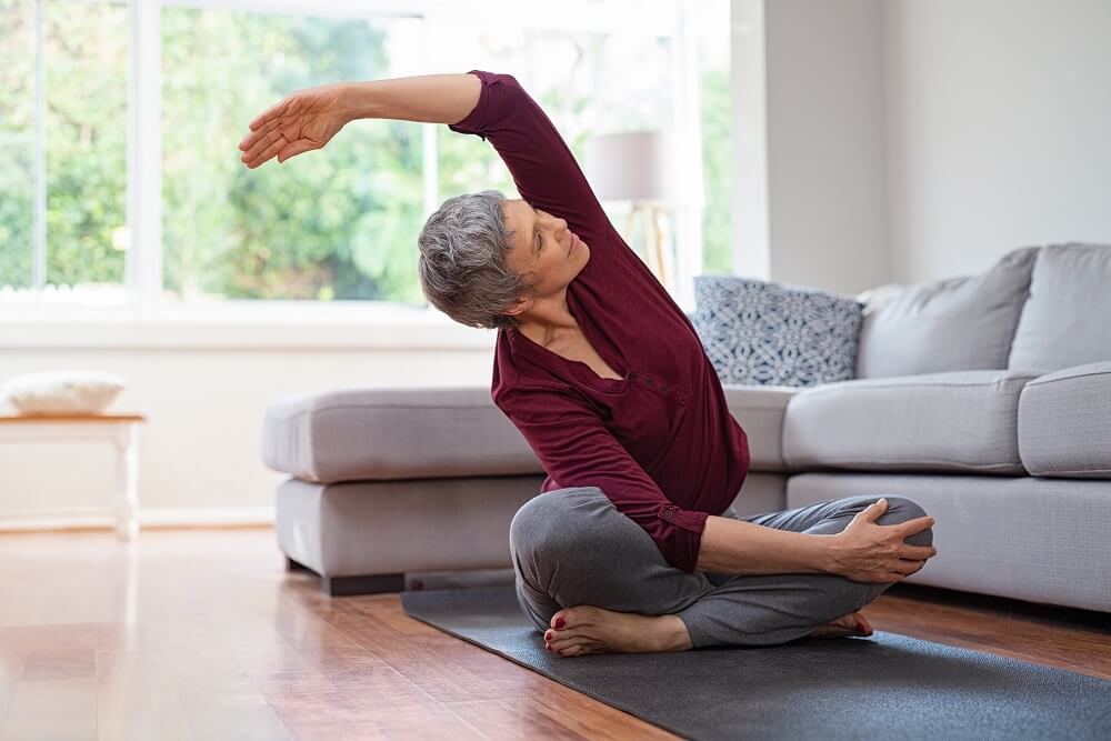 ältere Frau macht stretch-übungen im Schneidersitz auf dem Boden vor dem Sofa