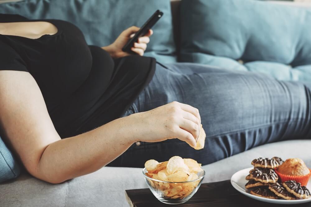 Übergewichtige Frau liegt auf Sofa und greift zu Chips und Süßigkeiten