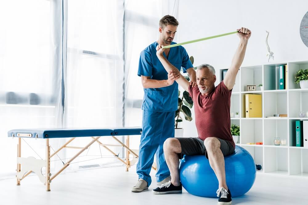 Älterer Mann macht Übungen auf einem Gymnastikball mit der Hilfe eines Physiotherapeuten 